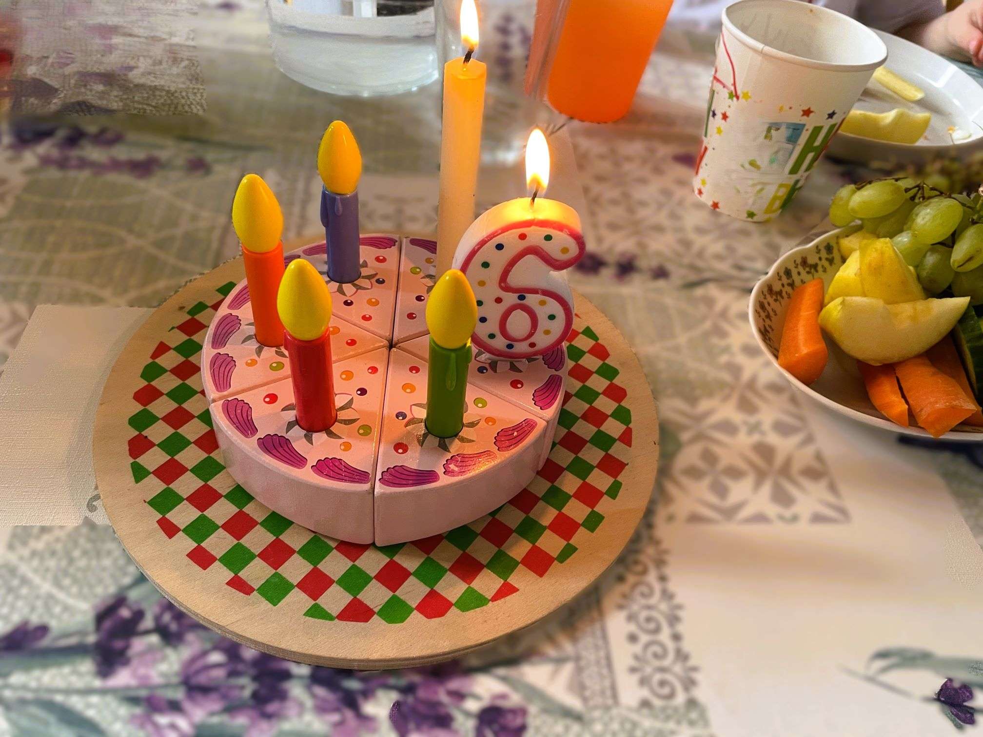 Auf dem gedeckten Tisch steht ein rosaner Holzkuchen mit vier Spielzeugkerzen und zwei echten Kerzen darauf, eine davon in Form einer 6. Es gibt keine feuerfeste Unterlage.
