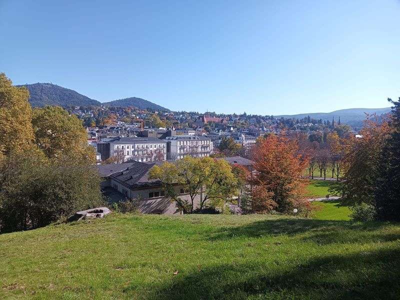 Der Blick auf eine herbstliche Landschaft mit grüner Wiese, bunten Bäumen und Städtchen.